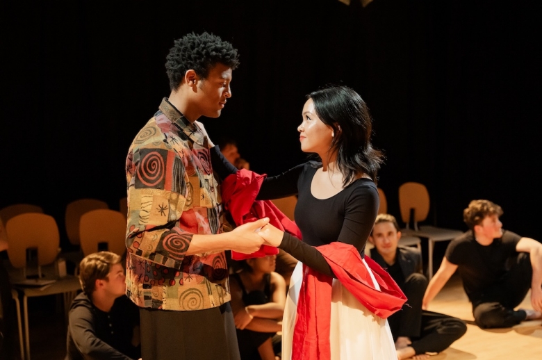 Two actors stand and face one another in mid-performance as other actors sit on the stage during the final presentation of The Winter's Tale.